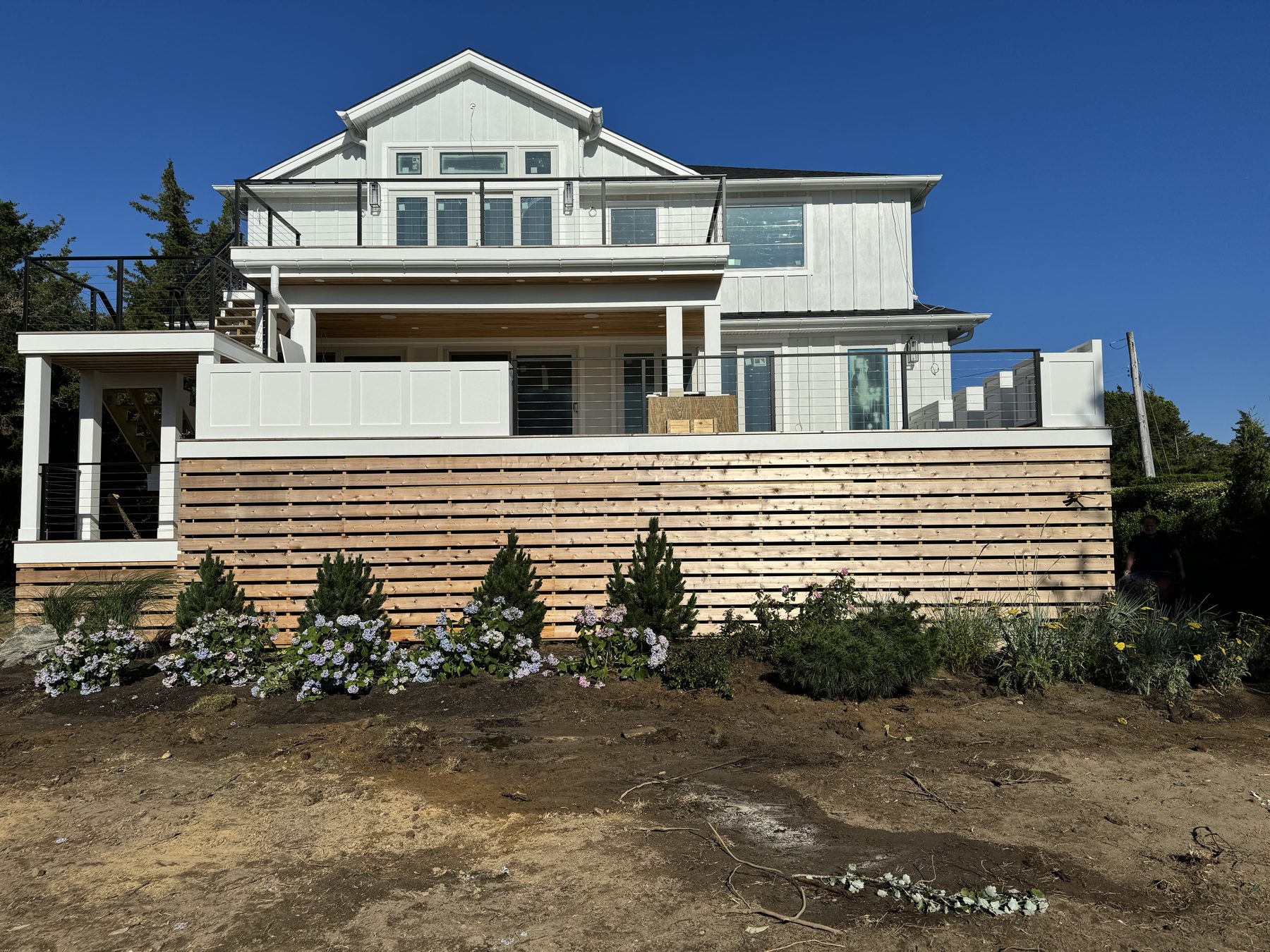 A modern white lakefront home with a long horizontal cedar-slat fence, freshly installed hydrangeas and shrubs along the foundation.