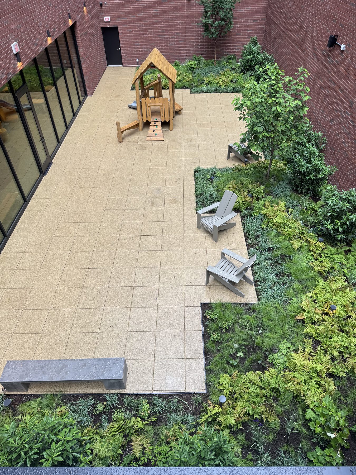 An overhead view of a stone courtyard patio with Adirondack chairs and a wooden arbor, surrounded by lush garden beds.